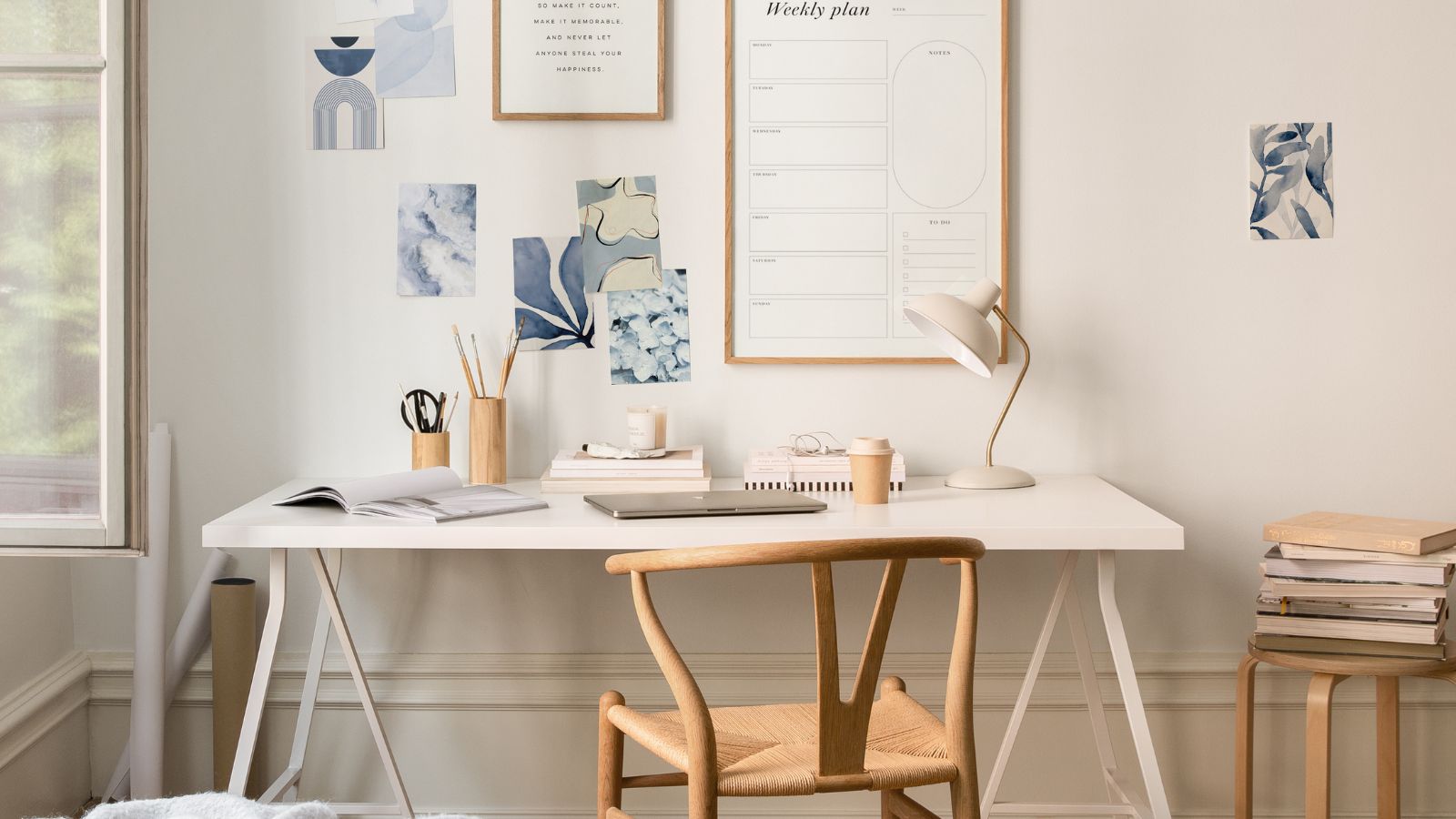 A minimalist white desk and wooden chair in a bedroom, with a stack of books on a stool next to it, ad blue pictures and a whiteboard on the wall above. 
