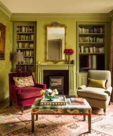 A lime green color drenched living room with contrasting patterned carpet and footstool, a burgundy velvet armchair with a matching beige upholstered one, a gold statement mirror, roses in a vase, and symmetrical color drenched bookshelves