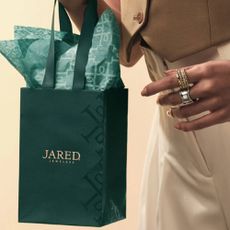 A person holding a small Jared gift bag, wearing a variety of gold and silver stacking rings from Jared.