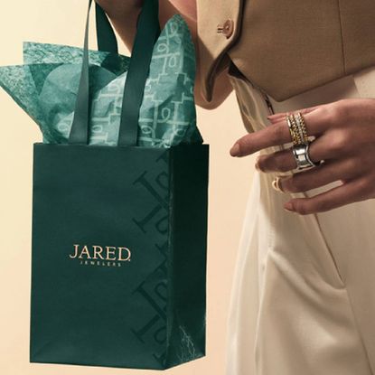 A person holding a small Jared gift bag, wearing a variety of gold and silver stacking rings from Jared.
