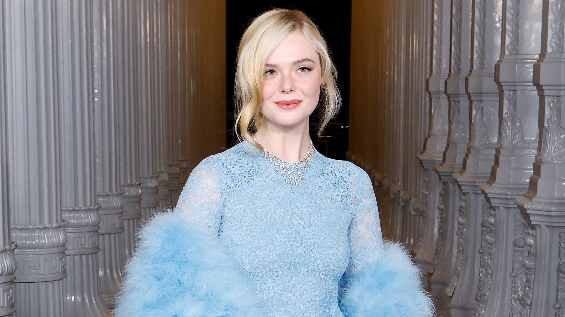 Elle Fanning wearing a blue sheer dress and a matching fur shawl. Her hair is in an updo and two curls are framing each side of her face. 