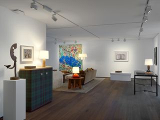 Art display room featuring white walls and ceilings, white ceiling lights and dark wood flooring. Wall art on display in different sizes. On the right is a black table with a lit up table lamp. On the left is a tartan designd cabinet rested against the white all with a lit up table lamp