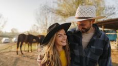 An attractive young couple hugs in front of horses on their ranch. They are wearing cowboy hats.