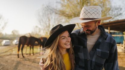 An attractive young couple hugs in front of horses on their ranch. They are wearing cowboy hats.