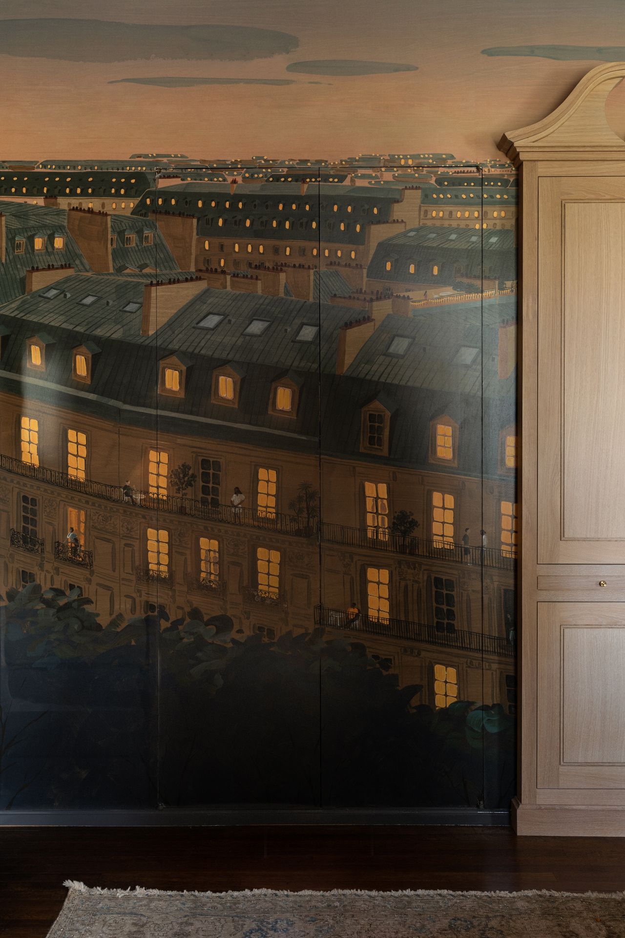 View of a wall painted in a Paris-theme city mural. The colors used are a dark tan-biege and a deep blue. The lights in the windows of the painting have a soft, warm glow as the painting is of the city in the evening. There is a wardrobe slightly in view