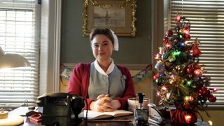 Megan Cusack in a nurse's outfit as Nancy sits at the surgery reception desk beside a Christmas tree in the Call the Midwife Christmas Special 2024.