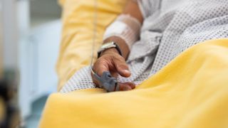 photo shows a close up of a man's hand laying relaxed on a hospital bed; a watch can be seen on his wrist and there's a blood pressure monitor on his pointer finger