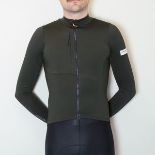 A white man in a dark green long sleeve cycling jersey and black cycling shorts stands against a white wall