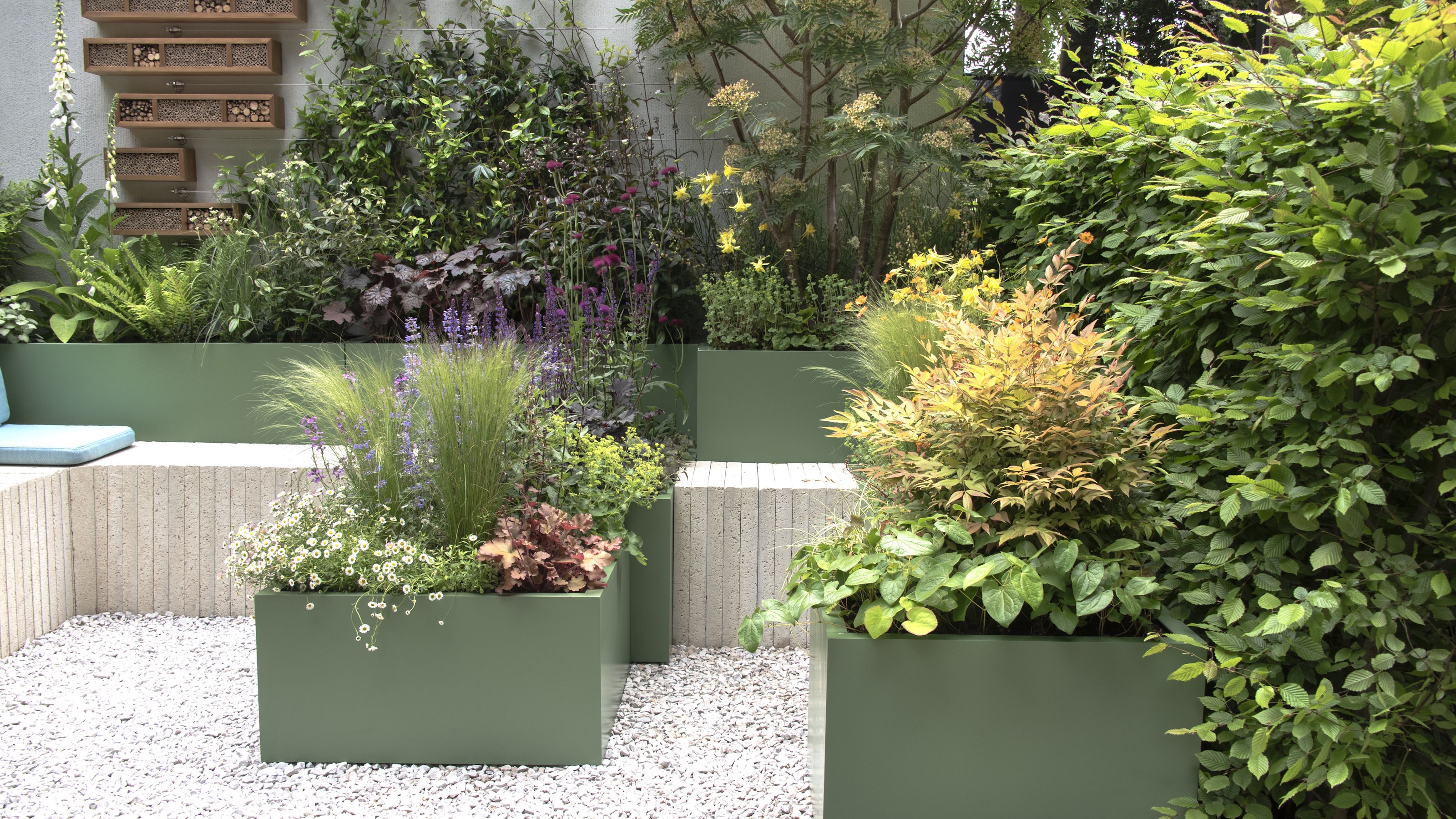 Plants growing in green containers and raised beds surround a seating area and gravelled patio area at the Chelsea Flower Show