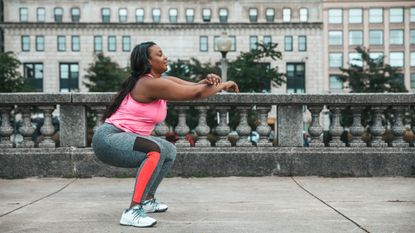 A woman in a sports vest, leggings and sneakers performs a squat on a sidewalk. Her knees are bent, hips pushed back and her arms are crossed in front of her. Behind her, we can see a big building with lots of windows and the tops of leafy trees.
