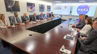 a group of people in business attire sit at a C-shaped table in front of a NASA logo
