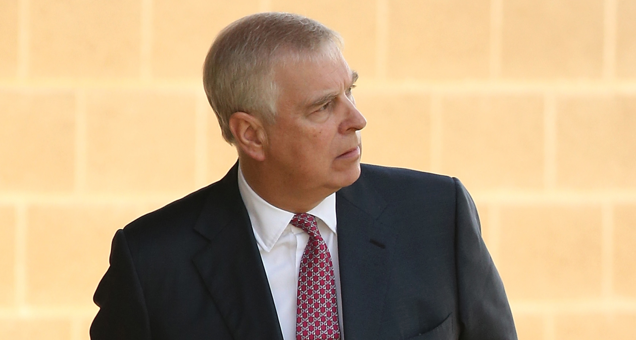 Ex-Prince Andrew wearing a red tie and suit standing in front of a yellow brick wall