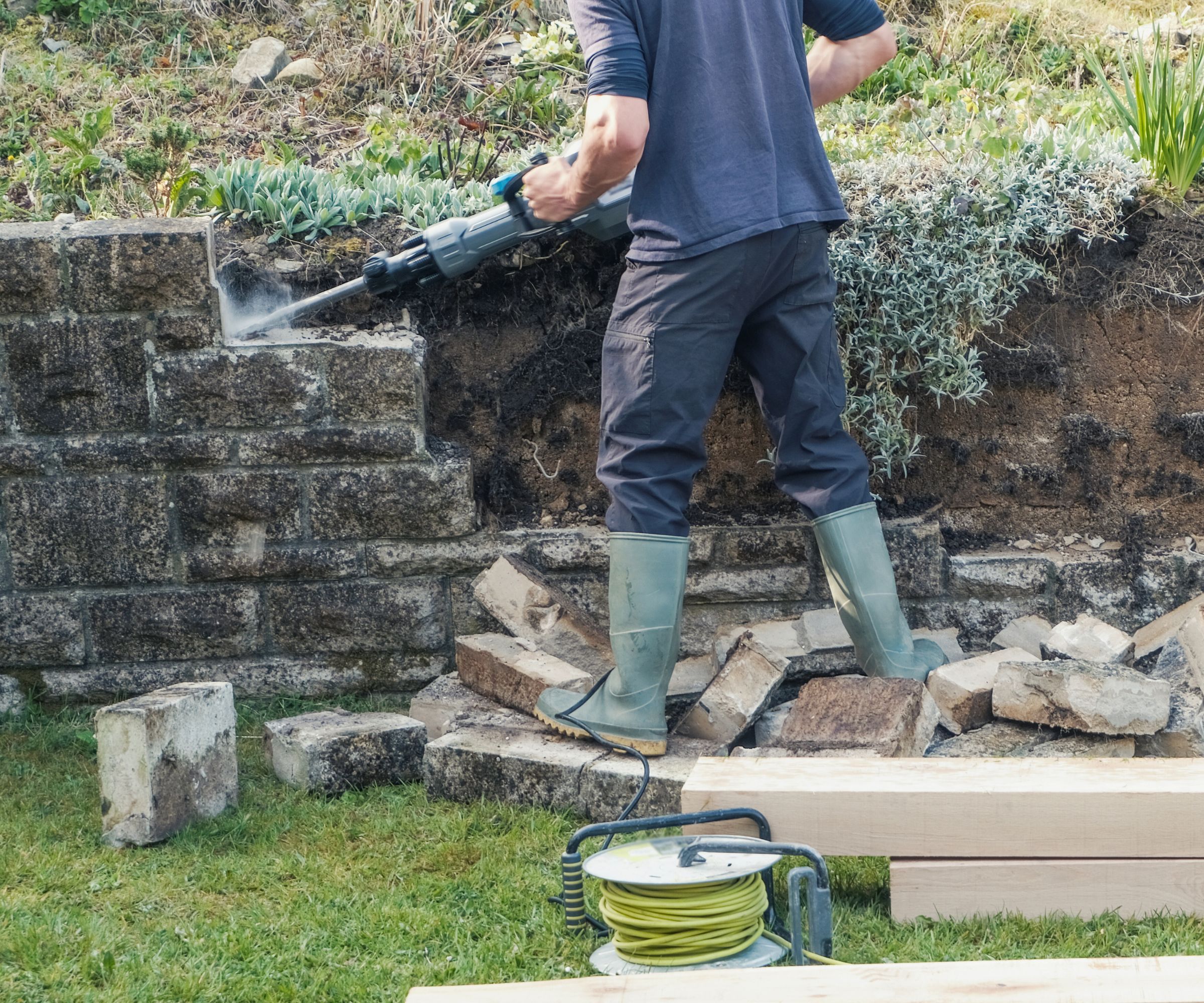 A man dismantling a retaining wall with a power tool surrounded by brick debris