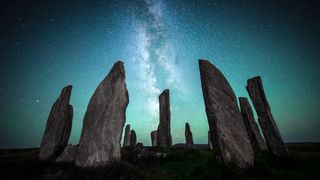 a view of a stone circle with several block-shaped stones reaching up to the sky. behind is the milky way, with a stone in front