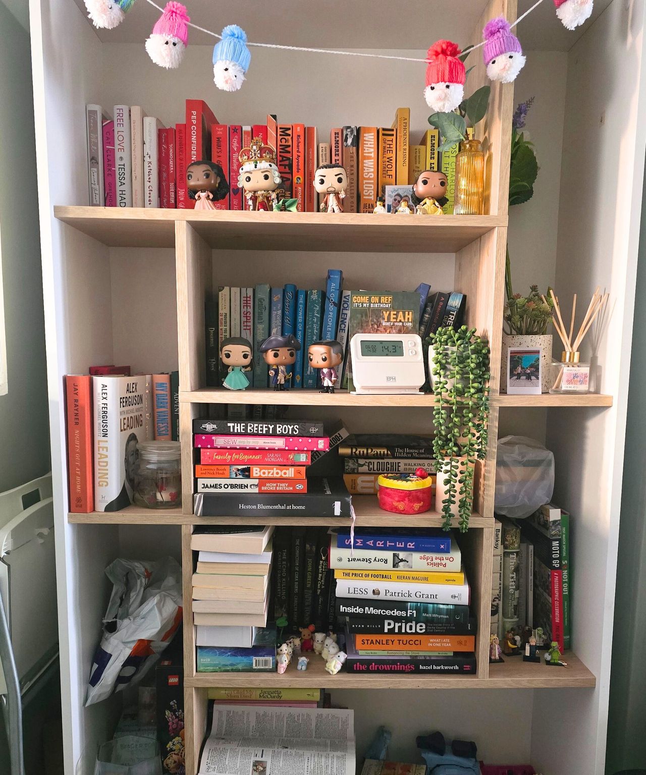 A white bookshelf with Santa decorations hanging across the top, and colorful books, faux plants, and Funko pops on it