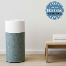 The Blueair Blue 3210 air purifier standing on a wooden floor next to a low wooden coffee table. Ideal Home approved logo placed in top right corner of image.