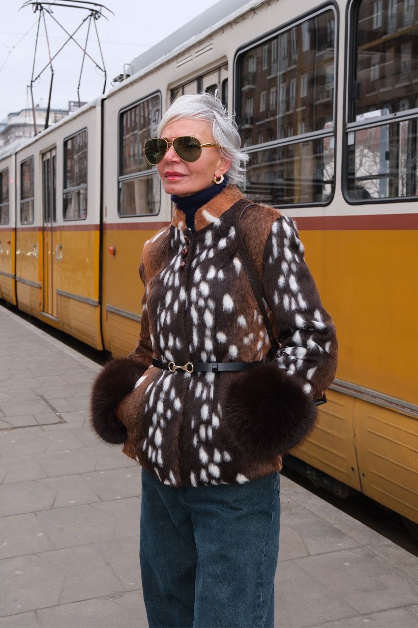 A woman standing outside in front of a yellow tram wearing a pony hair Bambi-print jacket styled with black turtleneck, baggy jeans, gold hoops, a black belt, and gold-frame aviator sunglasses.