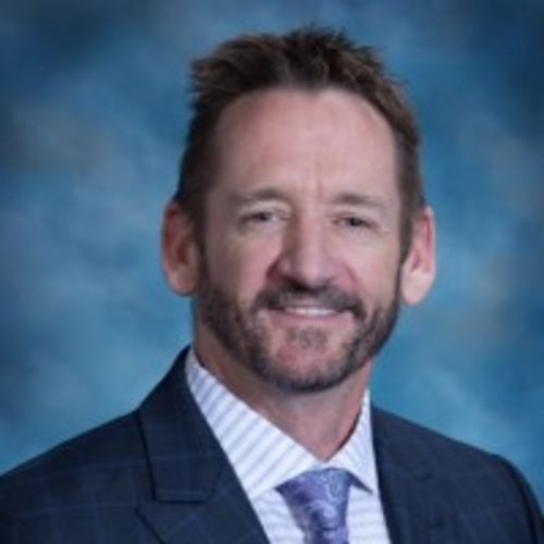Tom is a white man with dark brown hair and a beard, pictured in a navy suit against a mottled blue background