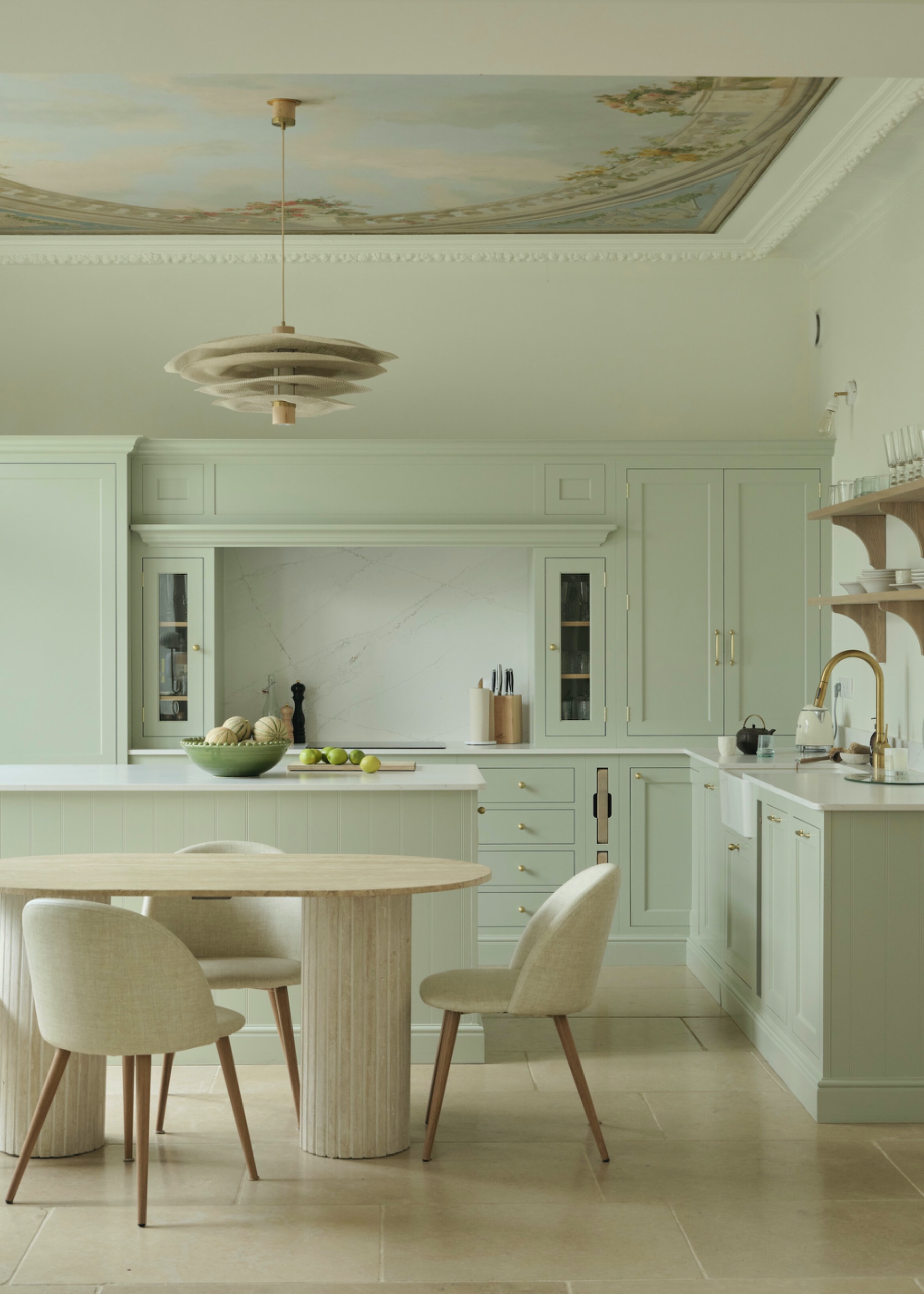 Pale sage-green kitchen cabinetry and island with brass hardware, stone flooring, timber dining table and a painted decorative ceiling.