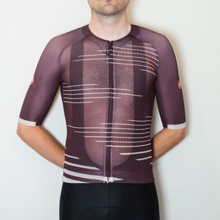 A white man in a purple cycling jersey and black cycling shorts stands against a white wall