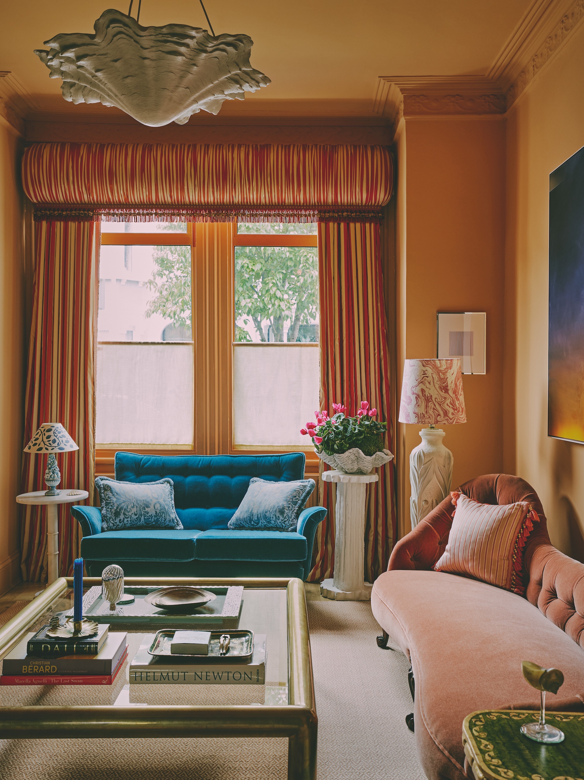 A traditional style living room with mustard yellow walls, and patterned curtains. There is a teal sofa in front of the windows on the far wall, with lamps either side and a peach chaise longue to the right and a glass coffee table in the middle of the space.