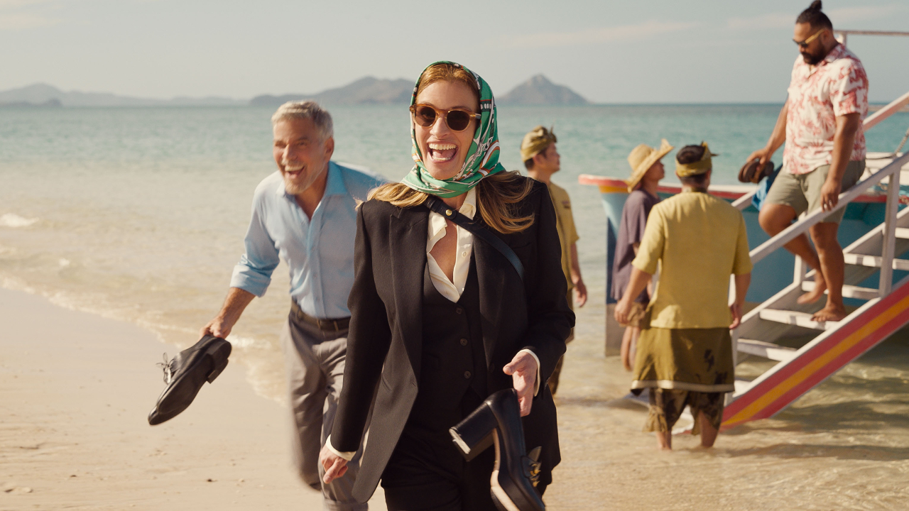 George Clooney and Julia Roberts excitedly walking on a beach, while holding their shoes in Ticket To Paradise.