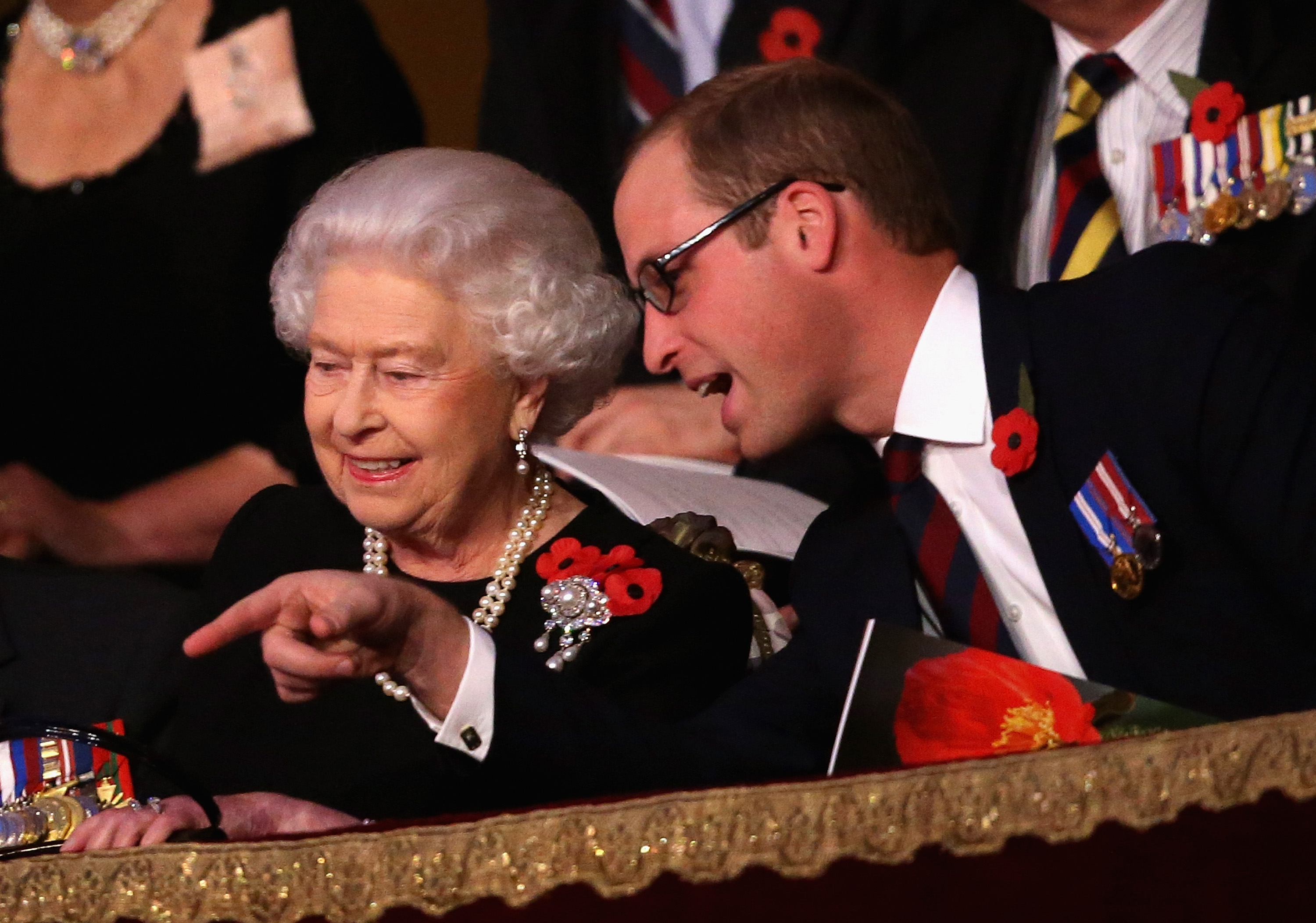 Queen Elizabeth wearing a black dress and pearls sitting next to Prince William, who is pointing at something and smiling
