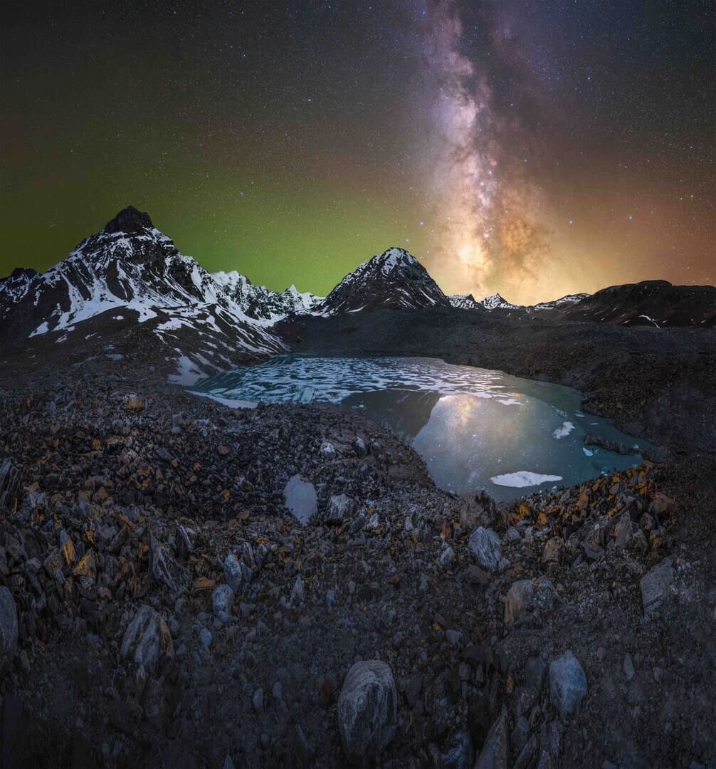 The Milky Way is visible in the night sky above an alpine lake with snow in the mountains