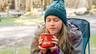 girl wearing beanie hat sitting in a camping chair near campfire holding a red enamel mug