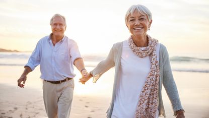 Portrait of a laughing senior woman leading her husband by the hand while walking on a sandy beach at sunset