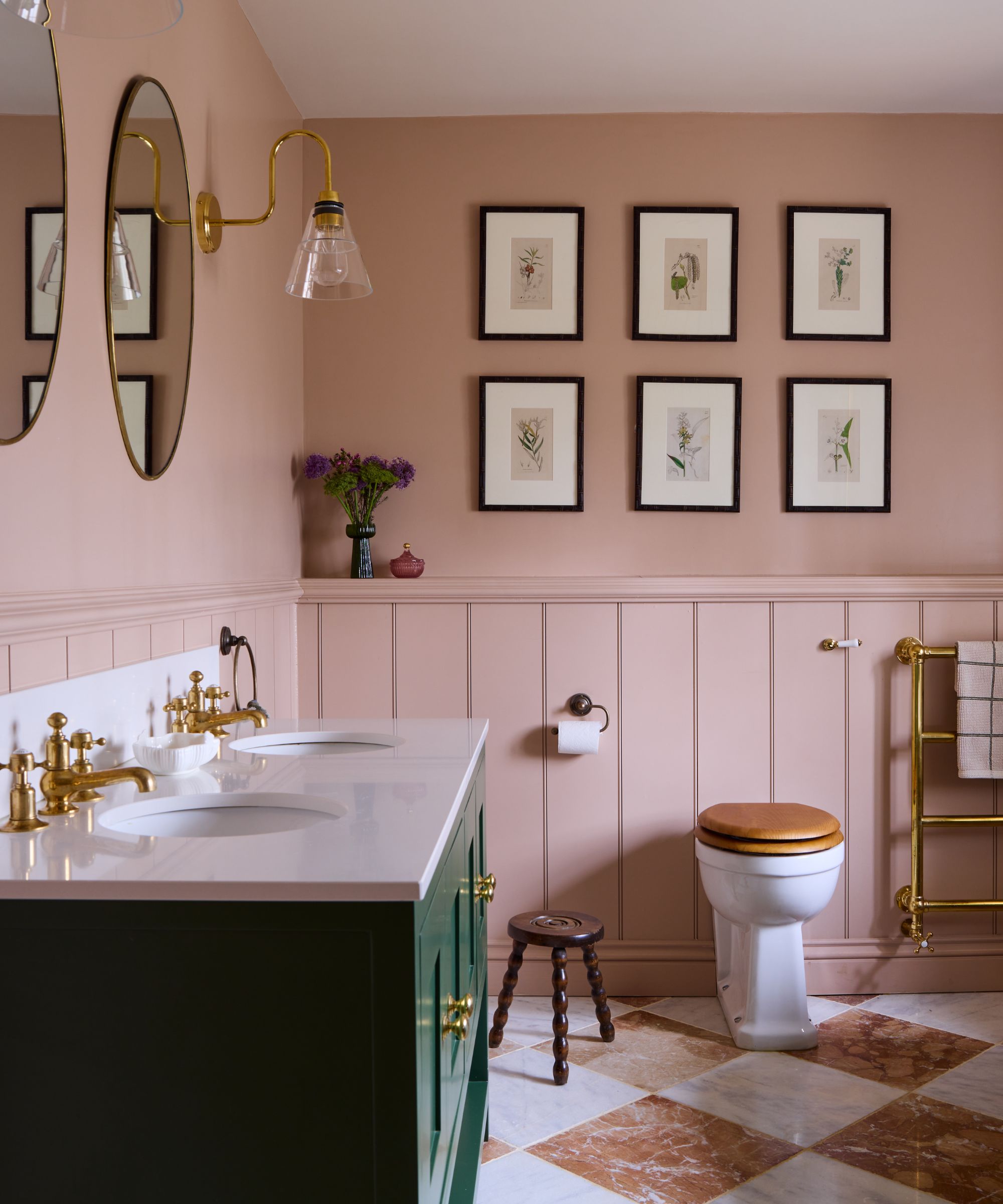 A traditional bathroom with plaster pink walls and panelling, a dark green vanity with double sinks, and six minimalist framed artworks on the wall.