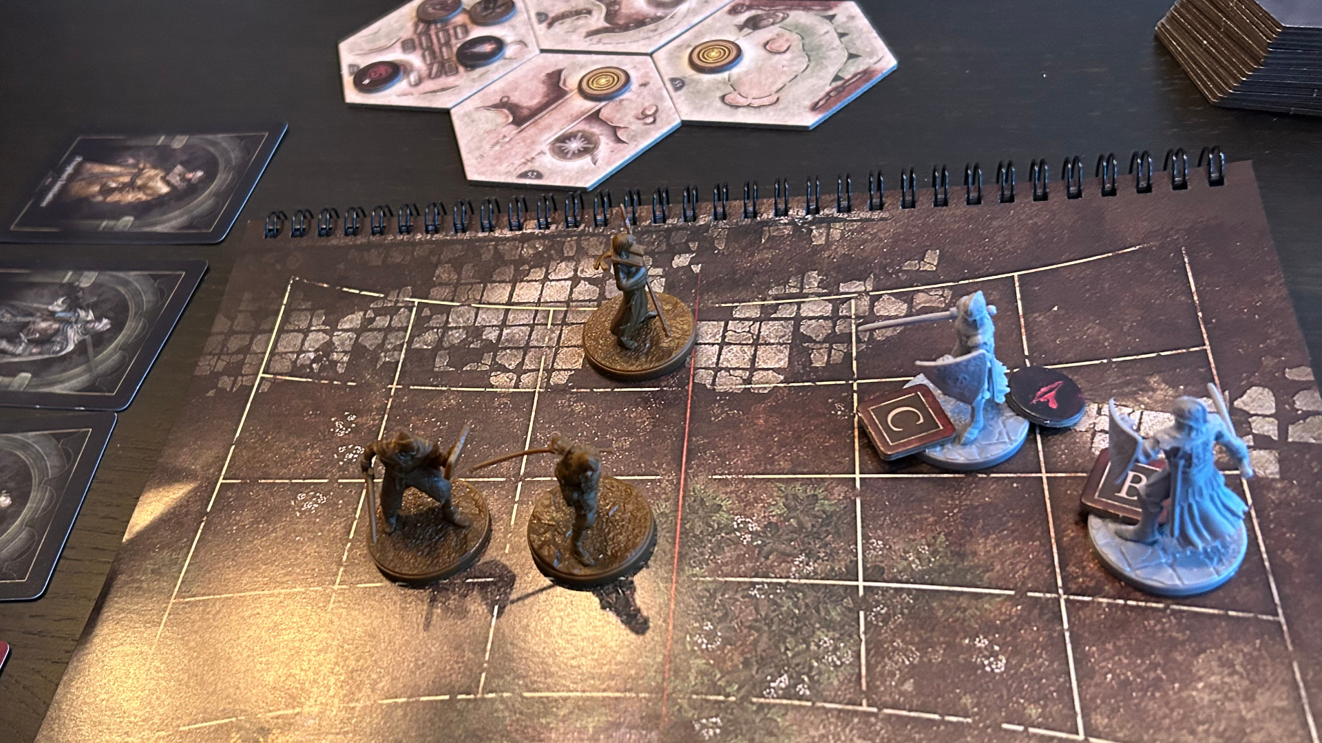 Elden Ring: The Board Game miniatures on an encounter book with a grid, beside board tiles and cards