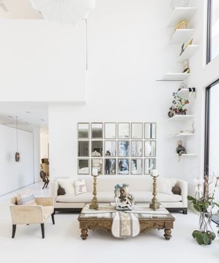 White living room with double height ceiling and shelves, sofa, armchair, coffee table, and gallery of mirrors on wall