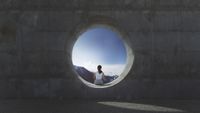 Photo looking at the back of a woman sitting alone in the opening of a concrete pipe on a sunny day. 