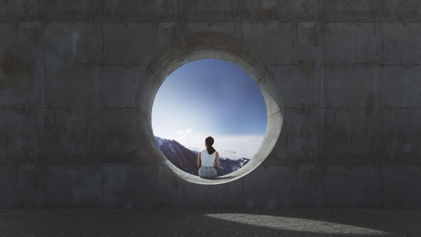 Photo looking at the back of a woman sitting alone in the opening of a concrete pipe on a sunny day. 