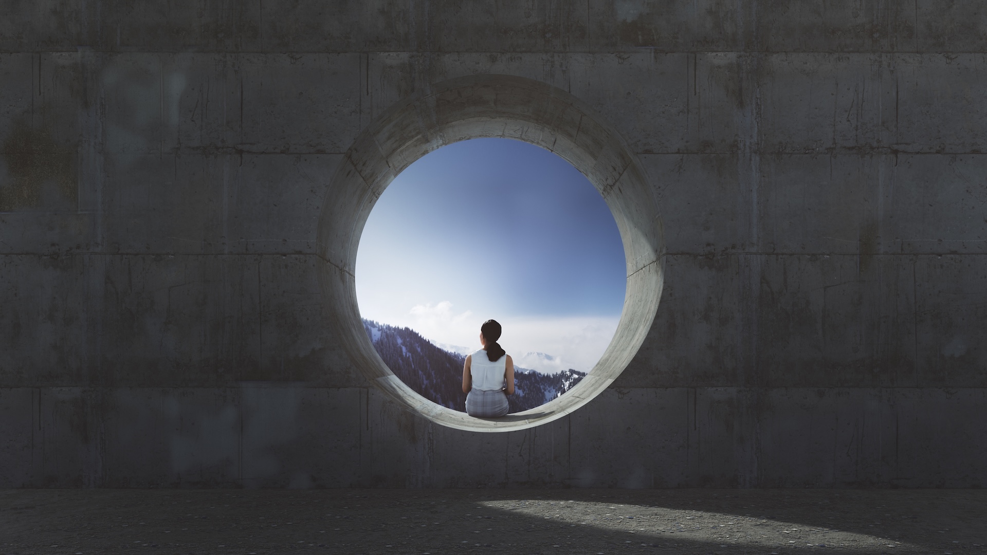 Photo looking at the back of a woman sitting alone in the opening of a concrete pipe on a sunny day. 