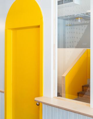 Interior view of a space at Les Fraiseurs featuring a white wall, a yellow door with an arched frame and a glass window offering a partial view of the staircase