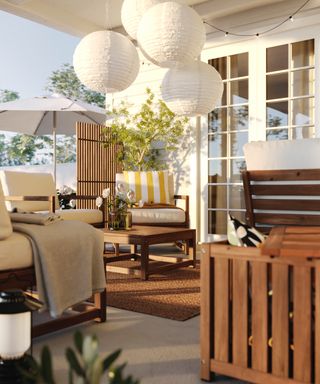 Paper style lanterns on a patio/porch with wooden cushioned seating, blankets, coffee table and jute rug, white parasol in rear