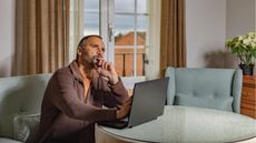 A man looks contemplative as he looks into the distance while sitting in front of his open laptop at a table.