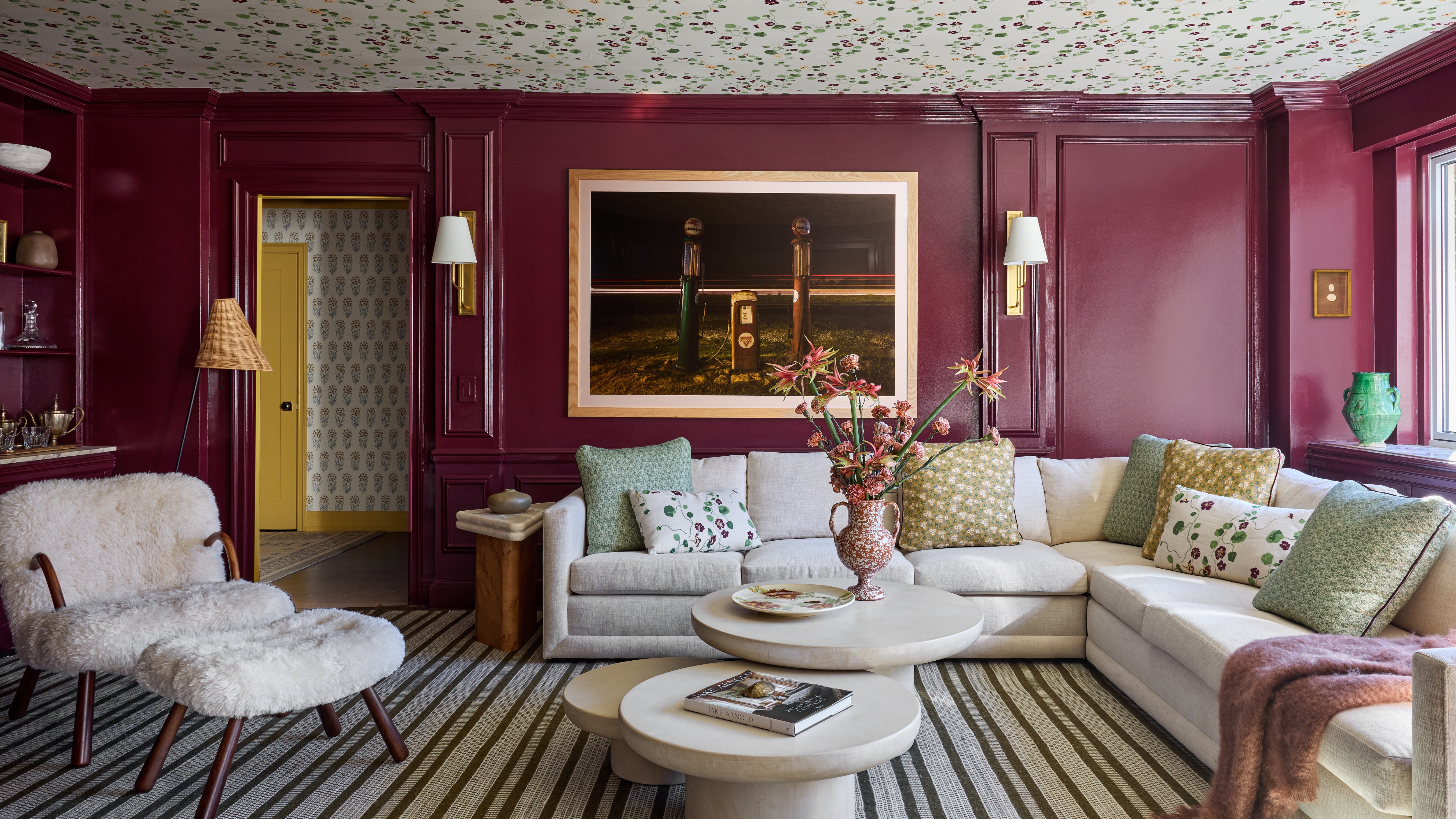 Moody den with plum-drenched walls, floral ceiling wallpaper, cream sectional, striped rug and contemporary artwork.