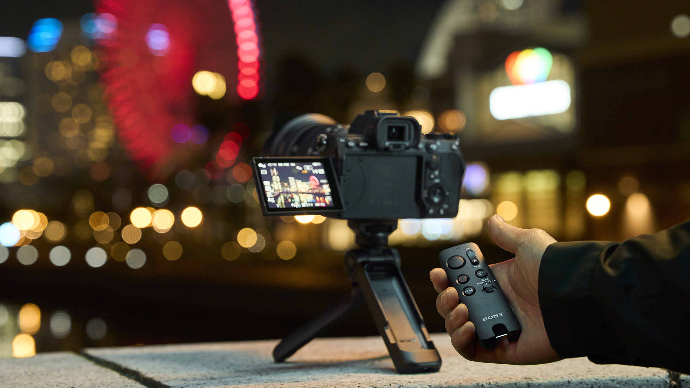 A person holds a Sony wireless remote control RMT-VP2 while adjusting a camera on a GP-VPT3 grip at night, with a colorful ferris wheel lit in the background