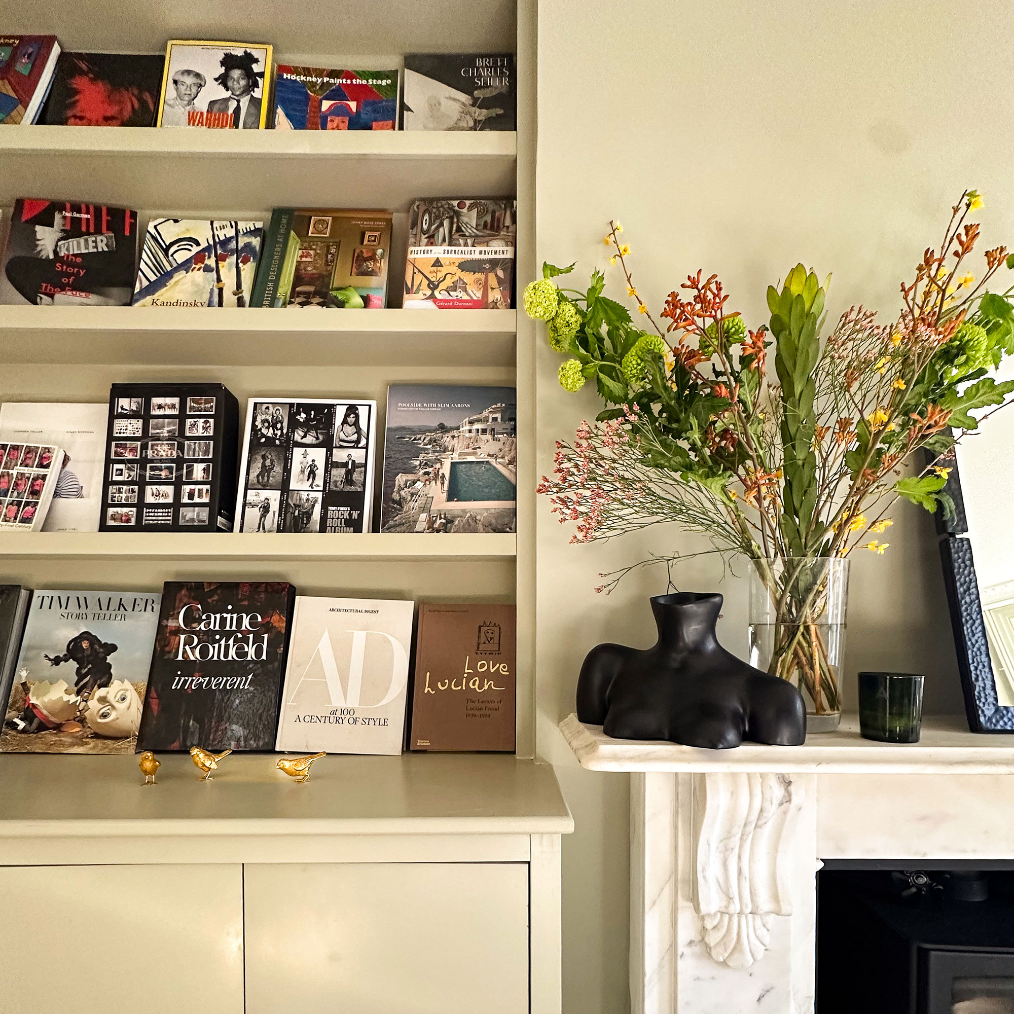 Pale green built-in bookshelves with coffee table books on display, beside marble fireplace mantlepiece with vase of flowers