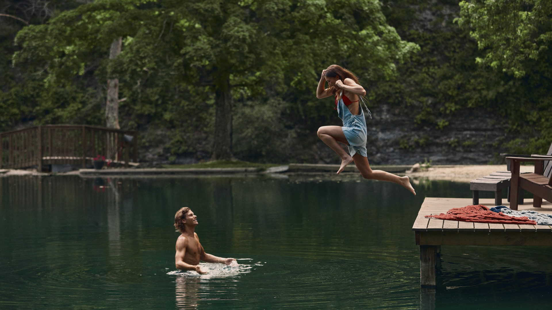 A person in a red bikini and blue overalls jumping off a dock into water while another person in the water looks on. The dock has a red and a blue blanket laying on it
