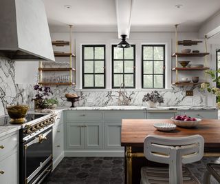 An L-shaped kitchen with a wooden kitchen island, light grey-green cabinets, marble countertops and backsplash, and a black and gold range cooker