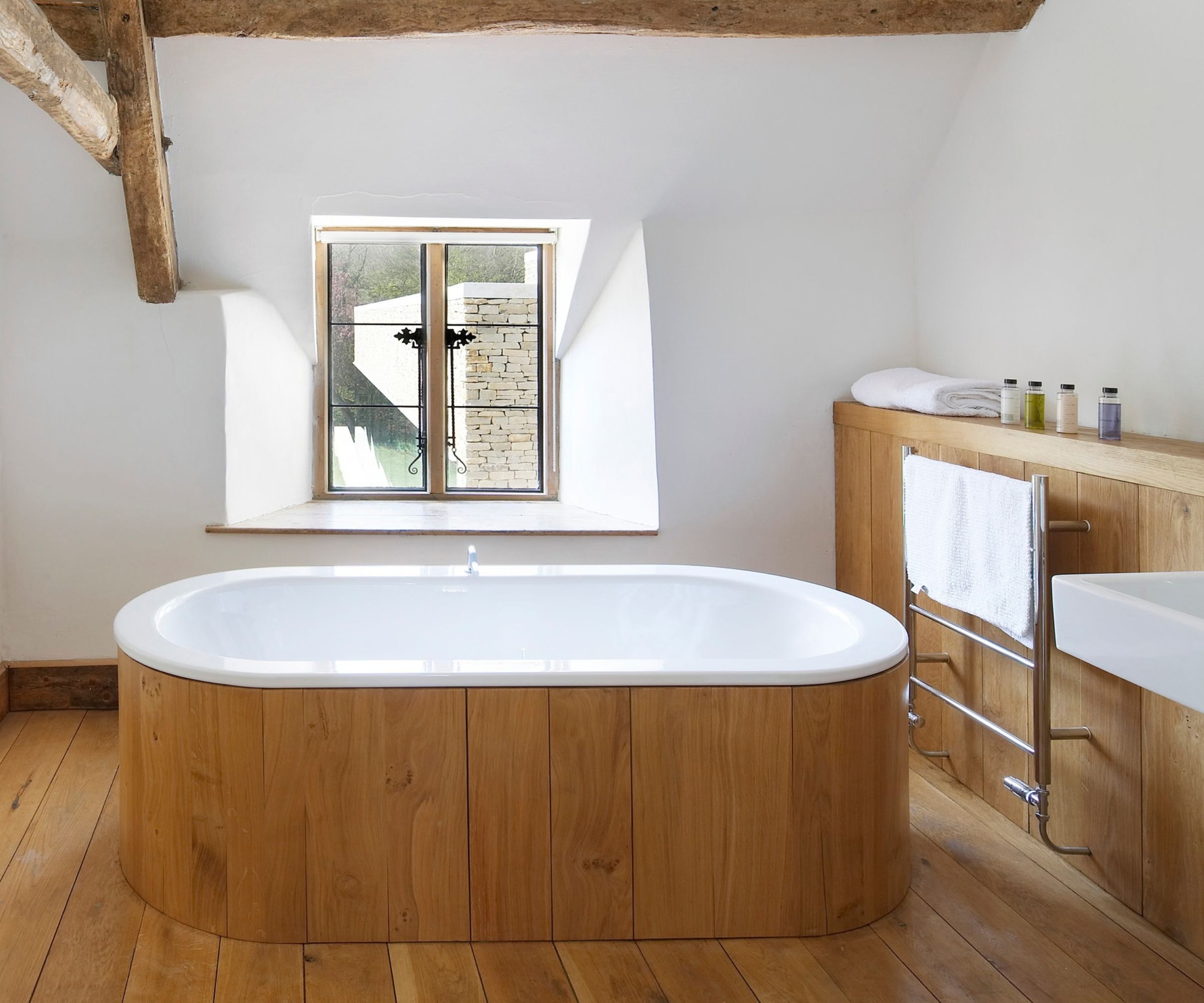 A characterful country bathroom with exposed oak ceiling beams and wide-plank oak flooring. A white oval bath with a curved oak panel surround sits centrally beneath a casement window, beside an oak-clad vanity unit with a chrome ladder towel rail and toiletries displayed on top.