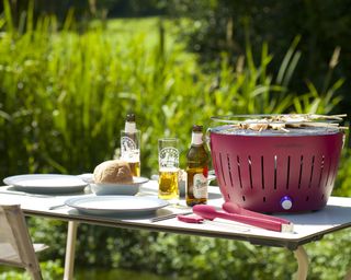 Red LotusGrill smokeless charcoal barbecue available from Cuckooland on outdoor table with beer and glasses with green grass in background