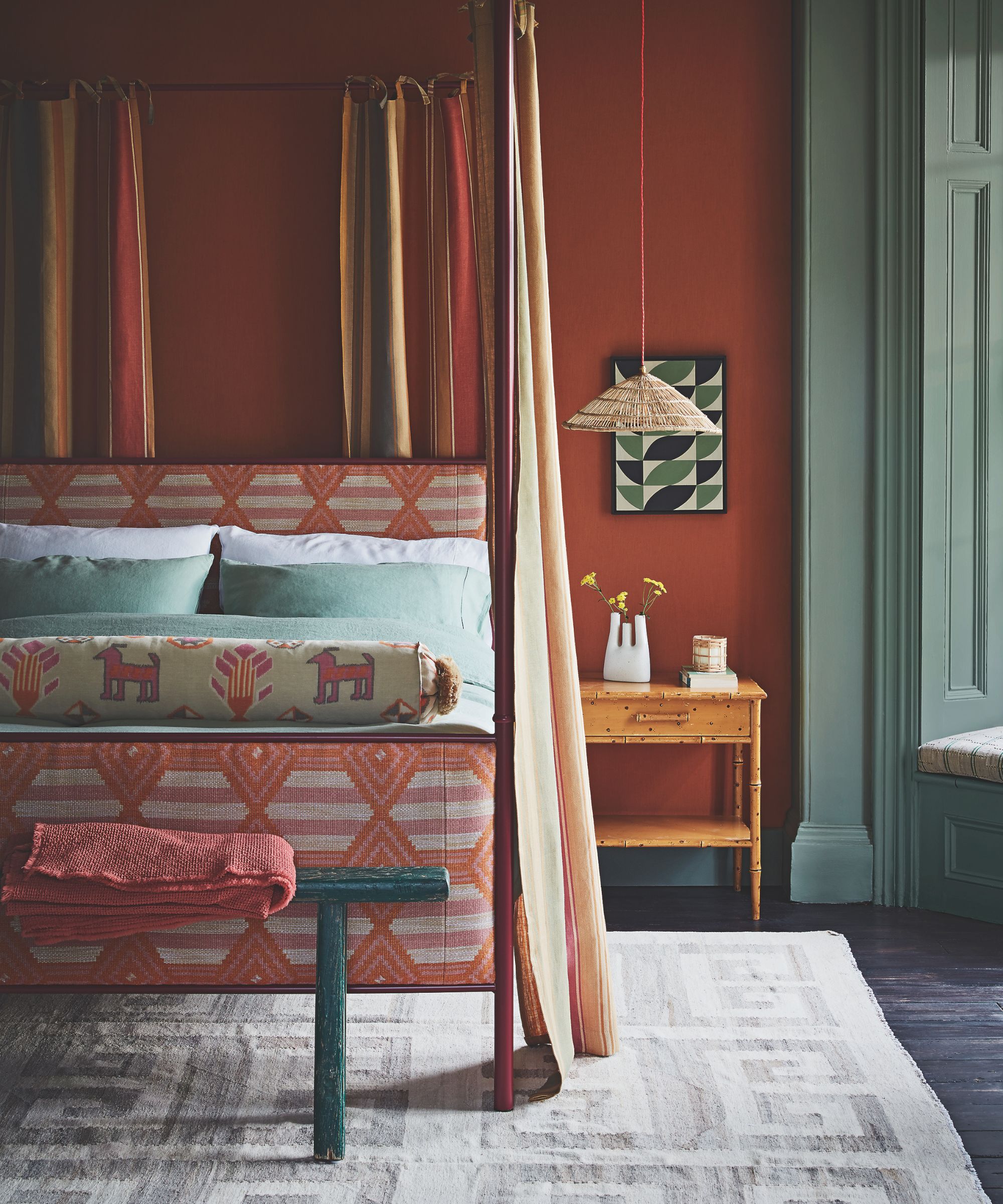 Warm bedroom with dark terracotta painted walls and four poster bed with flowing striped yellow and orange curtains. There is a geometric pattern on the headboard and foot board in dark orange, animal print pillows and sage green accents.