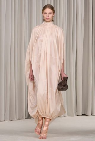 A snakeskin purse is shown in a photo of a model walking down the Ferragamo spring 2025 runway show, carrying a brown python clutch with a pink satin maxi dress and satin wrap up ankle flip flops