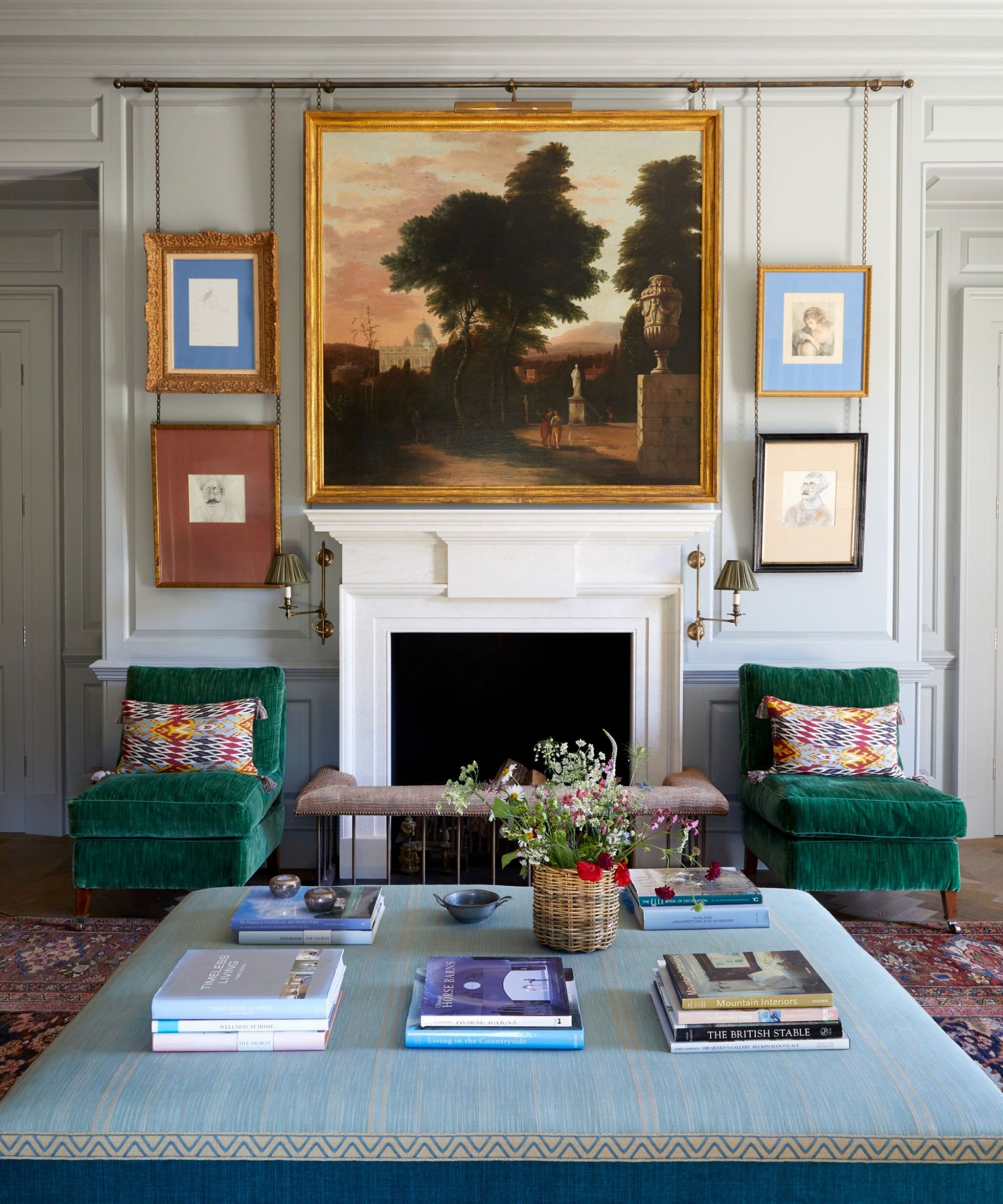 Living room with large vintage landscape above fireplace and smaller pictures on walls with two green velvet armchairs and large ottoman with books