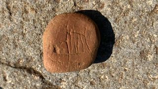 A 1-inch-long reddish clay stone has a carving of an incomplete Viking ship. The stone is photographed against a rock background.